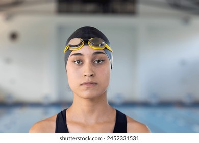 Biracial young female swimmer standing indoors in front of a pool, looking focused. She has brown skin, dark hair, and is wearing goggles on her head, unaltered. - Powered by Shutterstock