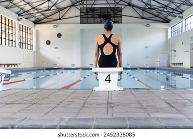 Biracial young female swimmer standing at pool edge indoors, preparing to dive, copy space. She has dark hair, wearing a black swimsuit, ready for training, unaltered - Powered by Shutterstock