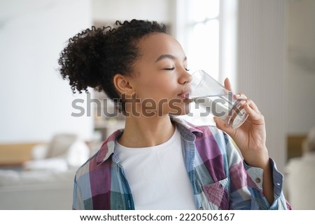 Biracial young girl drinks filtered clear water from glass, preventing skin dehydration. Biracial teen lady drinking mineral, caring about aqua balance in body, taking daily supplements at home.