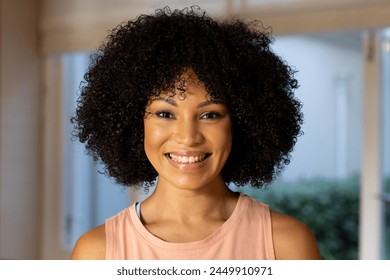 Biracial young woman with curly black hair smiling at camera at home. She wearing light makeup and simple pink top, unaltered - Powered by Shutterstock