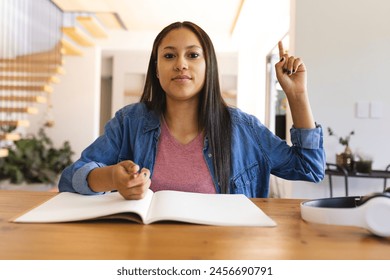 Biracial young woman holding pen during video call at home, wearing denim jacket. Sitting at table with long, straight brown hair, looking thoughtful, unaltered - Powered by Shutterstock