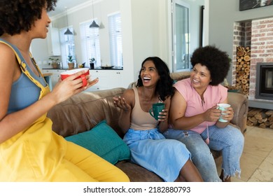 Biracial Young Woman Talking With Cheerful Female Friends Laughing While Sitting On Sofa At Home. Coffee, Unaltered, Friendship, Togetherness, Social Gathering, Enjoyment And Weekend Activities.