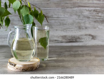 Birch sap in a transparent jug on a wooden stand. A tall glass of sap and a sprig of birch in the background. Light wooden background, space for text