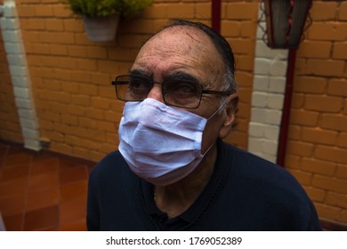 Bird Eye Angle Photography Of An Old Man (+70) With A White Face Mask, Glasses And A Blue Sweater Stand Up In The Backyard Looking At Camera. Profile Portrait