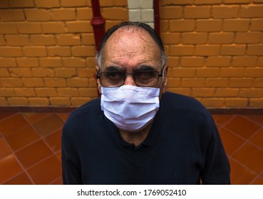 Bird Eye Angle Photography Of An Old Man (+70) With A White Face Mask, Glasses And A Blue Sweater Stand Up In The Backyard Looking At Camera. Profile Portrait