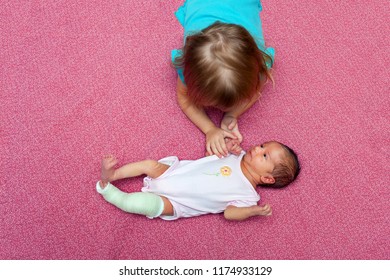 Bird Eye View Of An Older Sister Laying On Her Stomach, Holding The Hand Of Her Baby Sister To Comfort Her.  The Infant Has A Cast To Correct Her Congenital Club Foot.