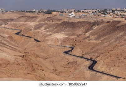Bird eye view on serpentine road 31 descending from city Arad to the Dead Sea, Judaean Desert, Israel. Panoramic landscape of the desert around the road. Cars on the road. City Arad on the background.