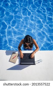 bird view of remote online working digital nomad woman in bikini with long black hair and laptop on a white table standing in a sunny blue water pool on Workation