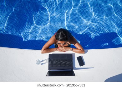 bird view of remote online working digital nomad woman in bikini with long black hair and laptop on a white table standing in a sunny blue water pool on Workation