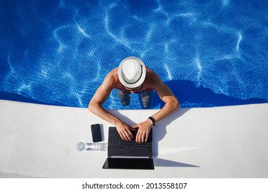 bird view of remote online working digital nomad man on workation with hat and laptop at a white table standing in a sunny turquoise water pool
