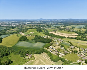 Birds eye perspective reveals patchwork fields, winding roads, clusters of houses, and green forests stretching across gentle hills beneath a clear blue sky