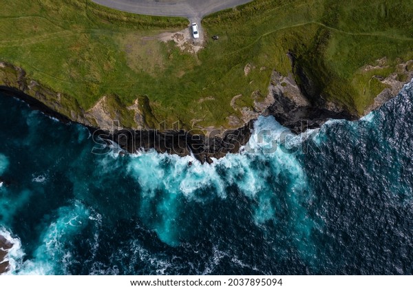 Birds eye\
view of car parked at end street of the famous Moher Cliff in\
Ireland with waves hitting the rocks during\
day