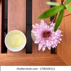 Birds Eye View Of Chinese Tea Served In A Procalain Ceramic Cup Next To Beautiful Pink Flower And Green Leaf On Top Of A Wooden Table.