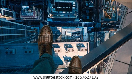 Bird's eye view from the clear glass deck on top of a high rise building. In the photo are the legs of a man in elegant shoes.