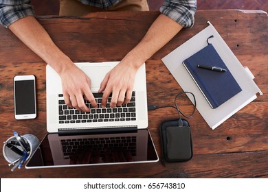 Birds Eye View Of Hands Typing On A Modern Notebook, Placed On A Durable Dark Wood Desk Along With Some Notes, Portable Storage, A Pen Holder And A Mobile Phone.