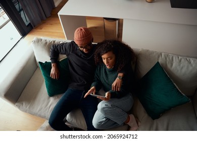 Birds Eye View Of Happy Multi-ethnic Couple Sitting On Sofa Using Electronic Tablet