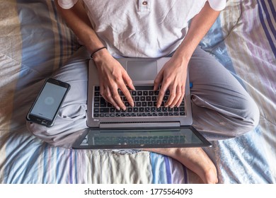 Birds Eye View Of A Young Man Working Studying In Home With Pajamas And Sitting On The Bed With Crossed Legs And Hands Tiping On A Keyboard. Laptop And Phone On His Legs. Blue/ Purple Striped Blanket