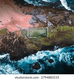 A bird's-eye drone shot of a large rectangular seaside swimming pool built into moss-covered rocky headland, adjacent to a pink-toned sandy beach with scattered sunbathers, surrounded by turbulent deep blue ocean waves.