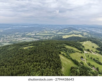 Bird's-eye view over a patchwork of forests, meadows, fields, and small clusters of houses with winding roads, distant town, and a cloudy sky