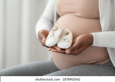 Birth Awaiting. Black expectant mother sitting with little baby shoes in hands, pregnant african american woman holding small sneakers near her belly, enjoying pregnancy time, closeup with copy space