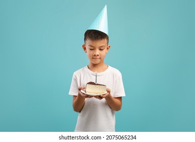 Birthday Boy. Happy Chinese Male Child Holding Piece Of Cake With Candle, Cute Little Asian Kid Wearing Party Hat Looking At Plate With Dessert, Enjoying Celebration, Standing On Blue Background