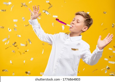 Birthday Boy Portrait. Fun Celebration. Amused Kid Blowing Party Horn In Confetti Rain Isolated On Orange Background.