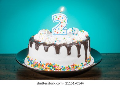 A Birthday Cake Bears A Lit Candle In The Shape Of The Number 2.