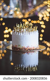 Birthday cake with candles, garland with bright bokeh lights on the background. The white cake is decorated with colored sprinkles and stands on a reflective surface. - Shutterstock ID 2008320836