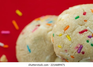 Birthday Cake Cookies: Two Vanilla Cookies With Sprinkles And White Chocolate Chips Baked In. Red Background. One Cookie In Close-up, One Blurred. Crumb And Loose Sprinkles To The Left Of Cookies. 