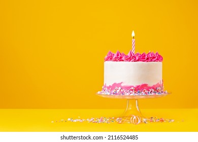 Birthday Cake with pink buttercream icing, colorful sprinkles and lit birthday candle over a bright and saturated yellow background. - Shutterstock ID 2116524485