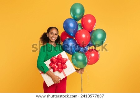 Birthday celebration. Cheerful african american teenager girl holds bunch of bright colorful balloons and gift box with ribbon bow, posing on yellow studio background, smiling to camera