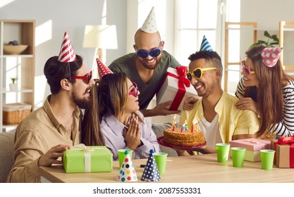 Birthday Celebration. Friends Congratulate Man On His Birthday And Give Him Birthday Cake With Candles. Birthday Celebration At Home. Party Hats On Head. Disposable Cups With Drinks And Gifts On Table