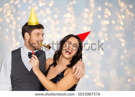birthday, celebration and holidays concept - happy couple with party blowers and caps having fun over festive lights background