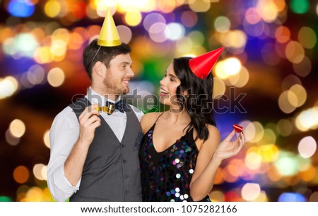 birthday, celebration and holidays concept - happy couple with party blowers and caps having fun over festive lights background