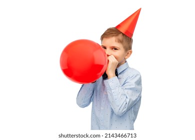 Birthday, Childhood And People Concept - Portrait Of Little Boy In Dress And Party Hat Blowing Balloon Over White Background