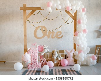 Birthday decorations with wooden arch, gifts, toys, balloons, garland and figure 1 for little baby party on a white wall background.