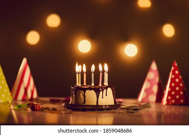 Birthday dessert concept. Drip cake with chocolate ganache and seven lit candles in centre of festive table with paper cone party hats against dark brown blurred background with bright bokeh lights - Powered by Shutterstock