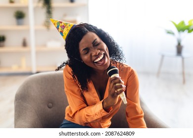 Birthday Fun. African American Woman Singing Song With Eyes Closed Holding Microphone Having Karaoke Party Sitting In Chair At Home, Wearing Festive Hat. B-Day Leisure Concept