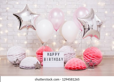 Birthday Party Concept - Close Up Of Air Balloons, Paper Balls And Lightbox With Happy Birtday Text Over Brick Wall With Lights