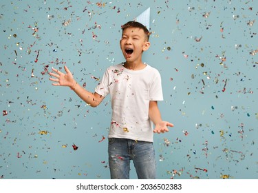 Birthday Party Concept. Happy Surprised Asian Boy Wearing B-Day Hat Standing Under Falling Silver Confetti, Joyful Emotional Male Preteen Kid Having Fun Over Blue Studio Background, Copy Space