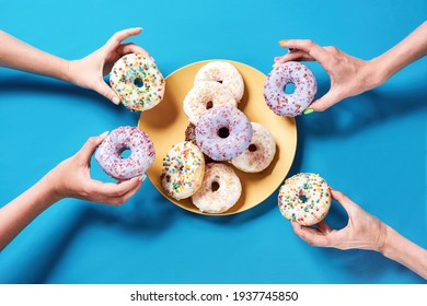 Birthday party. Different colourful round glazed donuts with sprinkles on yellow plate over blue background, four female hands taking donuts, top view. Unhealthy eating, desserts and junk food concept