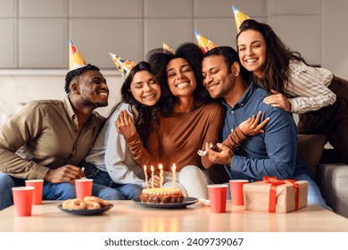 Birthday party. Joyful multicultural friends hugging bday lady celebrating and partying together, sitting near table with cake at modern home interior, wearing festive hats