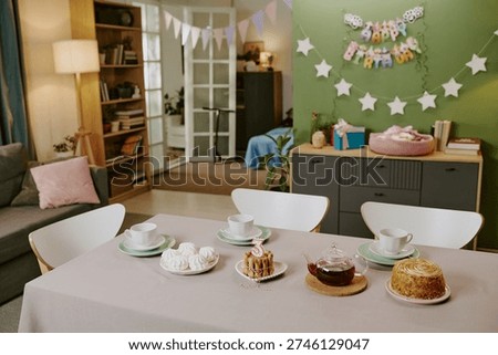 Birthday party table setting featuring cake with number three candle, pastries, teacups, and plates arranged in foreground, festive decorations and gifts visible in background