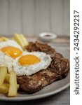 Bistec a lo pobre. Beef, eggs and fried potatoes on a plate on a wooden table with cutlery. Vertical close-up. Typical Chilean food
