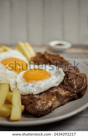 Bistec a lo pobre. Beef, eggs and fried potatoes on a plate on a wooden table with cutlery. Vertical close-up. Typical Chilean food