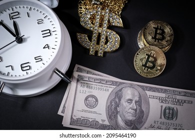 Bitcoin Coins Lie Next To A Stack Of $100 Bills, A White Alarm Clock And A Diamond-studded Dollar Chain Pendant