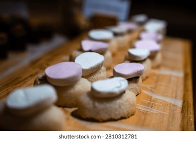 Bite sized dessert canapes sit in neat rows on a wooden board in the Maldives. Warm moody light and bokeh show soft bases with white and blush pink tops.