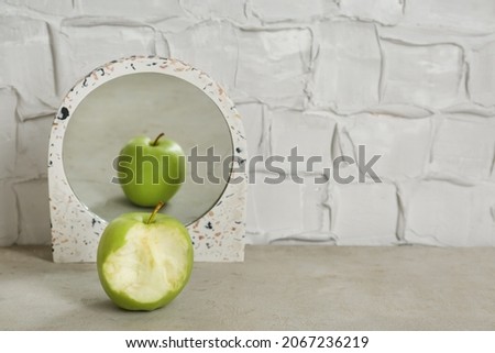 Bitten green apple near mirror with reflection of whole fruit on table, space for text