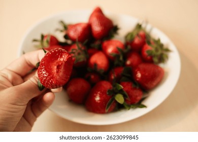 Una fresa mordida sostenida en la mano sobre un tazón blanco lleno de fresas frescas, resaltando la textura jugosa, el color rojo vibrante y la frescura de la fruta. Vista en primera persona