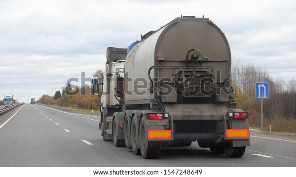 Bitumen truck\
with a round barrel semi trailer moving on a two-lane asphalted\
country road in autumn day, Logistics, hot liquid material road\
transportation, rear\
view